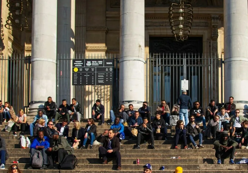 Esta imagen captura a un grupo diverso de personas descansando en las escalinatas de un edificio de arquitectura neoclásica, posiblemente un museo o institución cultural.