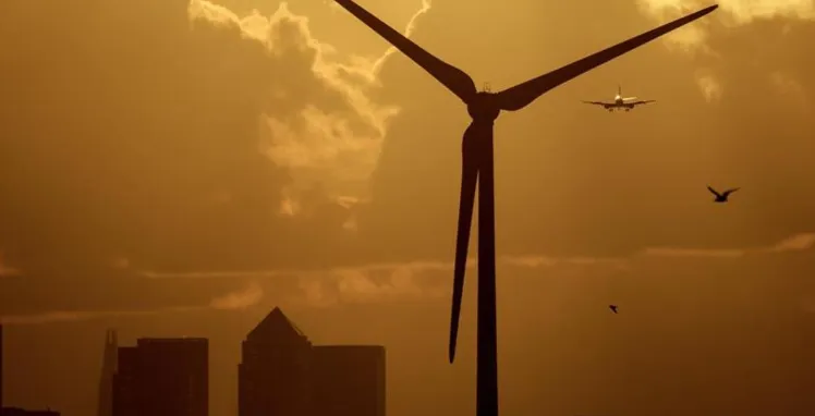 La imagen muestra un aerogenerador o turbina eólica en funcionamiento cerca de una zona urbana.