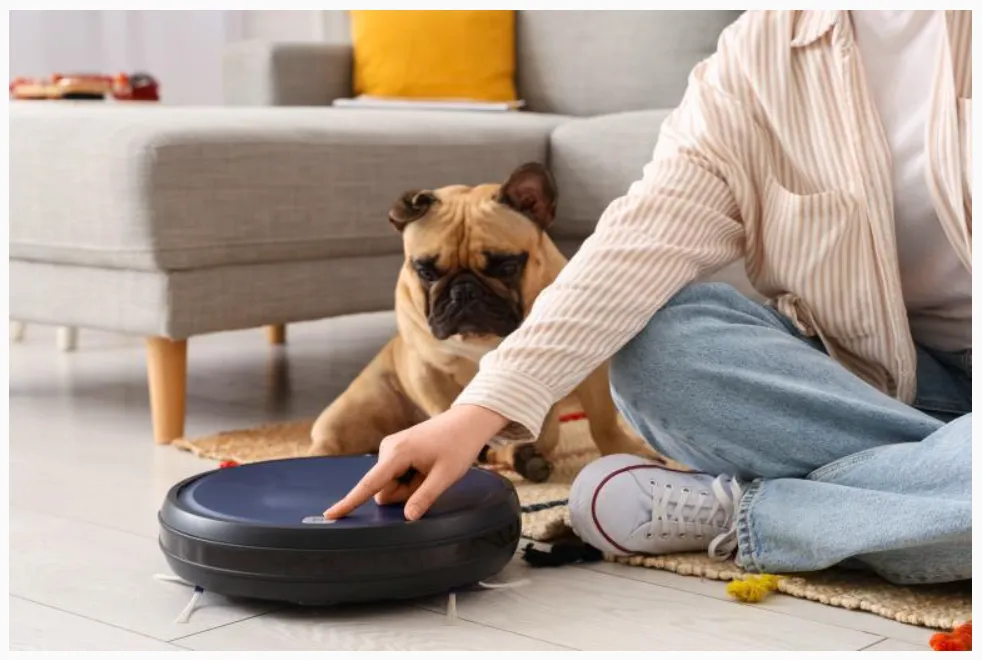 La imagen muestra una escena doméstica relajada donde una persona interactúa con una aspiradora robot, mientras un perro observa con curiosidad.