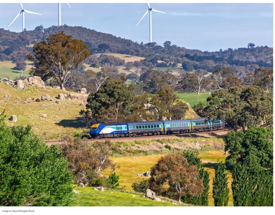 La imagen muestra el tren circulando por un paisaje rural montañoso cerca de Goulburn, Nueva Gales del Sur, acompañado por aerogeneradores. Es un tren de pasajeros XPT (Express Passenger Train) de NSW TrainLink operando en Australia.