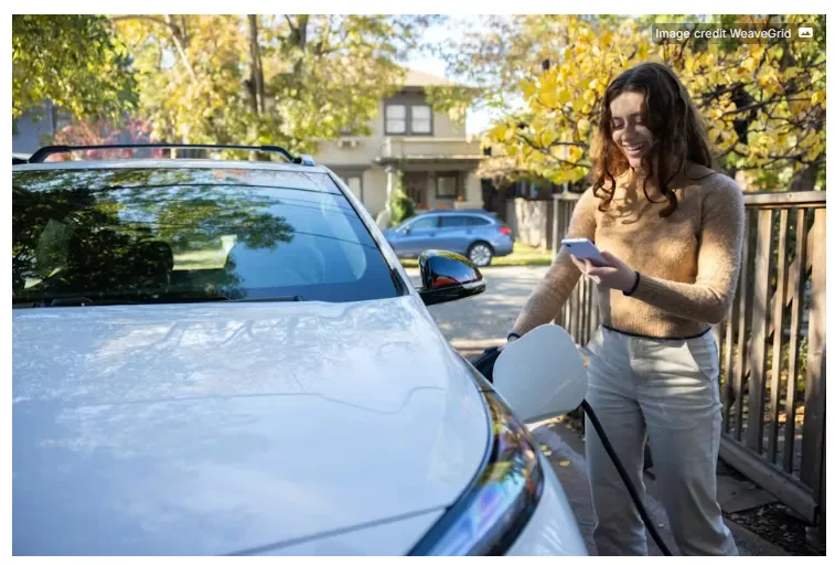 Esta imagen muestra a una persona cargando un vehículo eléctrico de color blanco mientras revisa su teléfono inteligente.
