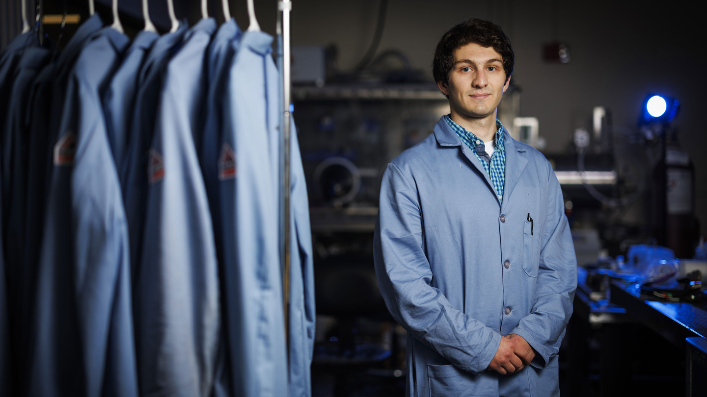 Joven investigador vistiendo una bata azul dentro de un laboratorio científico, posando junto a un perchero con más batas de laboratorio.