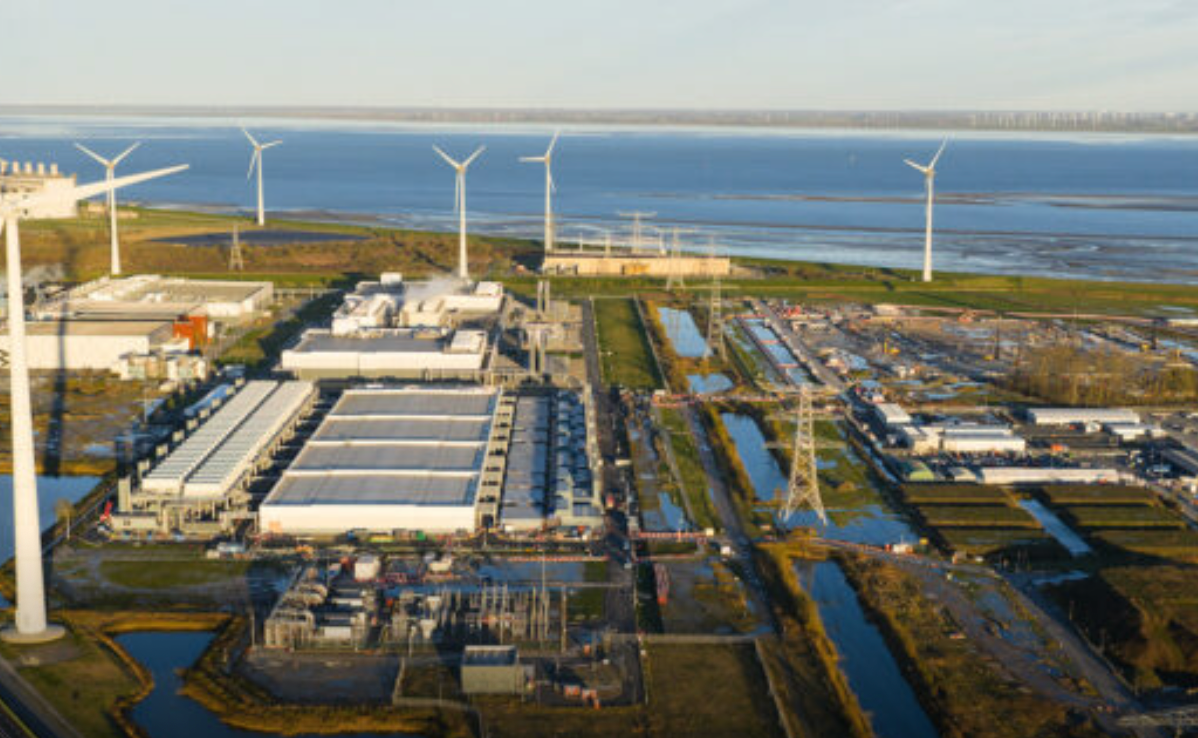 Vista aérea de una instalación industrial y de infraestructura energética situada junto a la costa, rodeada de aerogeneradores y torres de alta tensión.